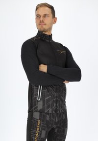 Schwarze Sportjacke mit hohem Kragen, verziert mit goldenen Akzenten und einem geometrischen Muster. Passende schwarze Leggings mit ähnlichen Designs.