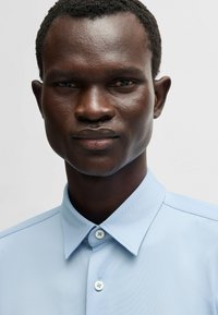 Chemise à col bleu clair avec un tissu texturé. Présente un devant à boutons avec deux boutons visibles et un design de col pointu.