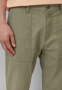 Khaki broek met een gestructureerde stof, zijzakken, rechte pasvorm en sluiting met bovenste knop, met een hand op de tailleband geplaatst.
