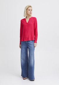Långärmad röd blus med V-ringning och rynkade axeldetaljer, tillsammans med vida blå denimjeans. Slitstyg, avslappnad passform.