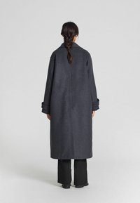 Lungo cappotto grigio in lana con una texture liscia, vestibilità rilassata e grandi bottoni. Presenta un colletto a punte e spalle basse.