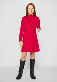 Piros, hosszú ujjú ruha magas nyakkal, csillogó derékrész részlettel és bővülő szoknyával, fekete térd fölé érő csizmával párosítva.