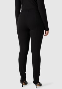 Pantaloni neri attillati realizzati in un materiale elasticizzato con una texture liscia. Dettagli delle cuciture notevoli che scorrono lungo i lati, abbinati a scarpe nere con tacco alto.
