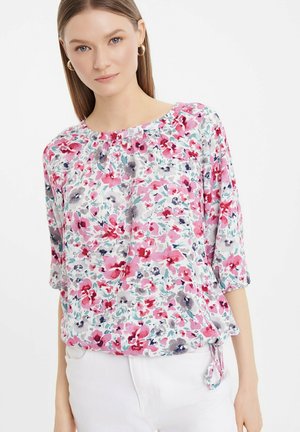 Bloem blouse met een ronde halslijn, halve mouwen en een strikdetail aan de onderkant. Kleuren zijn onder andere roze, paars en blauw op een lichte achtergrond.
