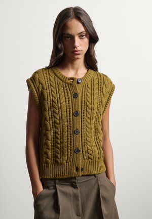 Giovane donna indossa un cardigan senza maniche color senape lavorato a trecce con bottoni neri e pantaloni verde oliva a vita alta, mani nelle tasche.