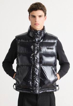 Junger Mann in glänzend schwarzer Pufferweste mit "HUGO"-Logo, Hände in den Taschen, schwarzes langärmeliges Shirt, vor weißem Hintergrund.