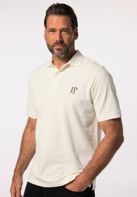 Cremefarvet polo-shirt lavet af tekstureret stof, med en krave med to knapper og et lille gråt logo på venstre bryst.