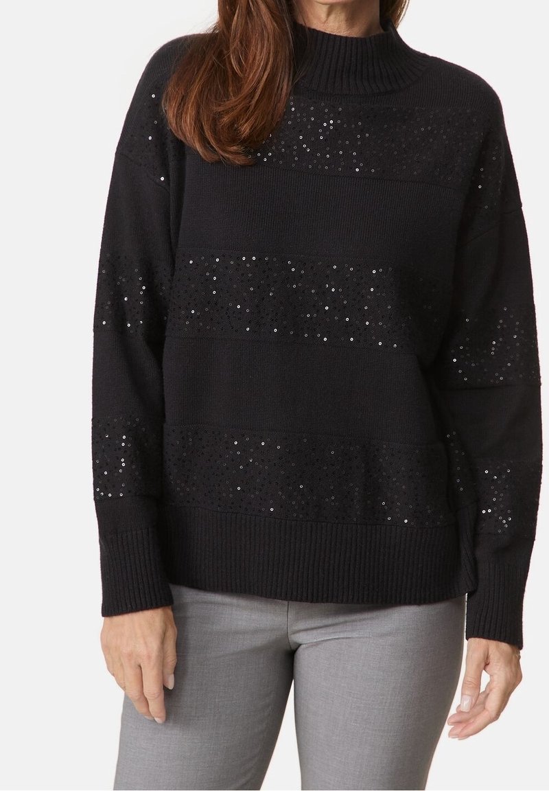 Femme portant un pull en maille noir avec des rayures horizontales en sequins, associée à un pantalon gris clair, sur un fond uni.