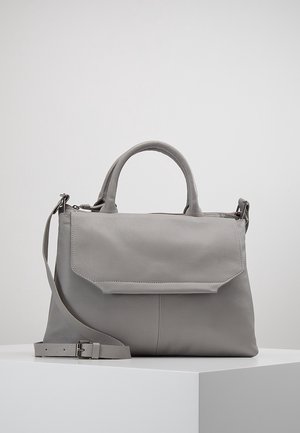 Sac à main en cuir gris avec rabat, deux petites poignées et une bandoulière réglable reposant sur un piédestal blanc.