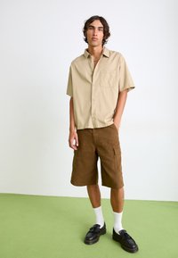 Beige short-sleeve shirt with a chest pocket, paired with brown cargo shorts. Black shoes with thick soles; white socks visible.