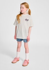 Vit bomull T-shirt med en blommig logotyp på bröstet, kombinerad med blå denimshorts och rosa sportskor med orange detaljer.