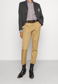 Grå kavaj med en enstaka emblem, randig skjorta, beige byxor och svarta ankelboots med spetsig tå och häl-detalj.