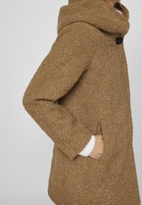 Abrigo de textura marrón con un gran cuello enrollado, bolsillos laterales y cierre de un solo botón. El tejido parece suave y cálido.