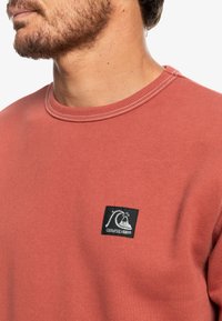 Rostfärgad sweatshirt med rund halsringning, med en strukturerad tygkvalitet. Svart fyrkantig lapp med vitt logodetalj på framsidan.