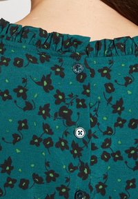 Turkoosi kukkakuosiset paita, jossa on rypytetty kaulus, musta kukkakuosi ja pyöreät napit takana kontrastivärillä.