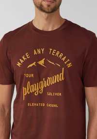 Bordowa koszulka bawełniana z żółtym nadrukiem przedstawiającym sylwetki gór oraz tekstem: "Make Any Terrain Your Playground", o luźnym kroju.