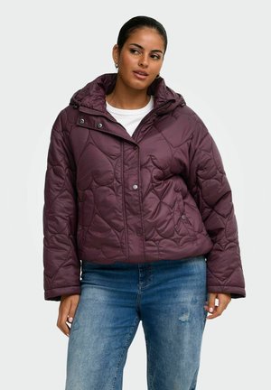 Frau trägt eine dunkelviolette gesteppte Jacke mit Kapuze und blaue Jeans, steht mit entspannten Händen an den Seiten.