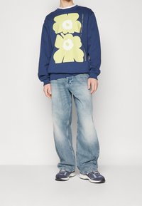 Námornická mikina s velkými žlutými květinovými potisky, kombinovaná se světle modrými volnými džíny a tmavými teniskami se šedými akcenty.