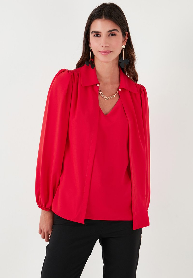 Red blouse with a collared neck, balloon sleeves, smooth texture, layered design, and v-neck. Worn over a coordinating top. Black pants.