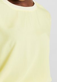 Nærbillede af en person iført en lysegul top med en cremefarvet ribbet rund halsudskæring på en ensfarvet baggrund.