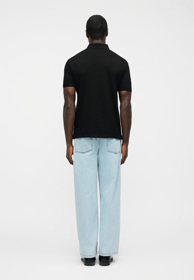 Άνδρας που στέκεται με την πλάτη στραμμένη, φορώντας μαύρο πόλο με κοντά μανίκια, ανοιχτό μπλε τζιν με ίσια πόδια και μαύρα παπούτσια σε λευκό φόντο.