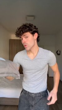 Giovane uomo con capelli ricci, indossa una maglietta grigia e jeans, tiene in mano un capo di abbigliamento piegato in una camera da letto con un letto e un armadio sullo sfondo.