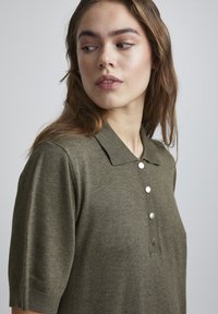 Polo vert olive en tissu texturé avec un col, des manches courtes et trois détails de boutons argentés à l'avant.
