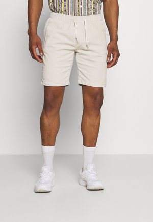 Homme portant un short beige avec cordon, des chaussettes blanches et des chaussures de sport blanches sur un fond uni.