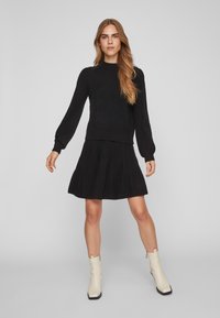 Schwarzer Strickpullover mit langen Puffärmeln und rundem Ausschnitt, kombiniert mit einem ausgestellten schwarzen Rock; dazu cremefarbene Ankle Boots getragen.