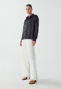 Camicia nera in satin con bottoni e colletto, abbinata a pantaloni bianchi a gamba larga e sandali neri con tacco. Tessuto morbido e vestibilità comoda.