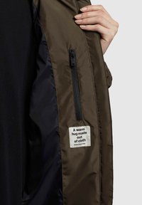 Olijfgroene jas met rits, interieur met zwarte voering. Bevat een label met de tekst "Een warme knuffel van stof" vlakbij de ritszak.