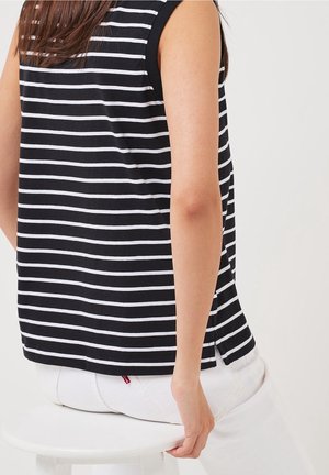 Femme assise sur un tabouret blanc, portant un haut sans manches à rayures horizontales noires et blanches et un pantalon blanc, la main posée sur le tabouret.