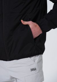 Schwarze leichte Jacke mit Rei�ßverschlusstaschen und elastischen Bündchen, neben weißen Shorts mit sichtbarem Logo, betont den Farbkontrast.