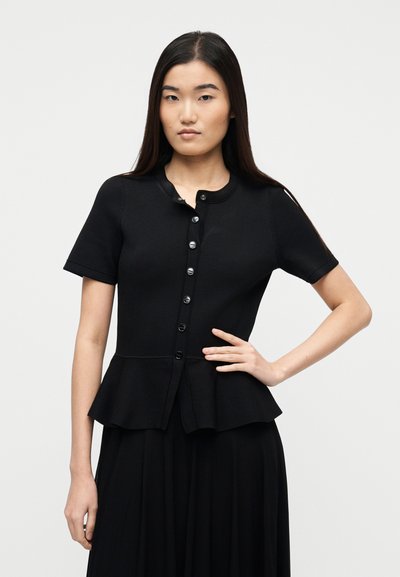 Νεαρή γυναίκα με μακριά σκούρα μαλλιά, φορώντας εφαρμοστό μαύρο κοντομάνικο πουκάμισο με κουμπιά και βολάν στη βάση, στέκεται με το ένα χέρι στη μέση της.