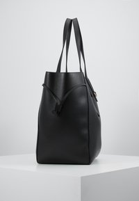 Czarna skórzana torba na ramię o fakturowanym wykończeniu, z dwiema długimi rączkami i zaciąganym zapięciem. Kwadratowa podstawa i zwężający się kształt u góry.