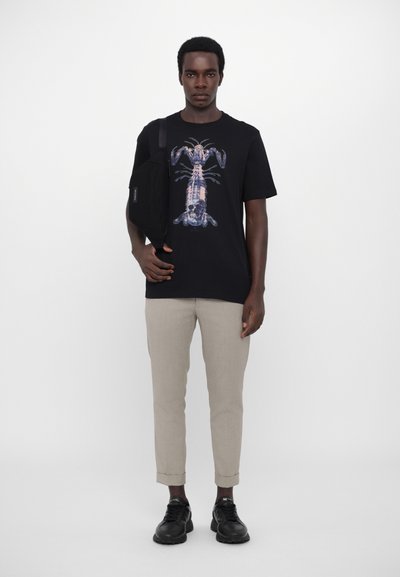 T-shirt noir avec un graphisme de homard, pantalon beige et baskets noires. Le mannequin tient un sac à bandoulière noir. Design simple et contemporain.