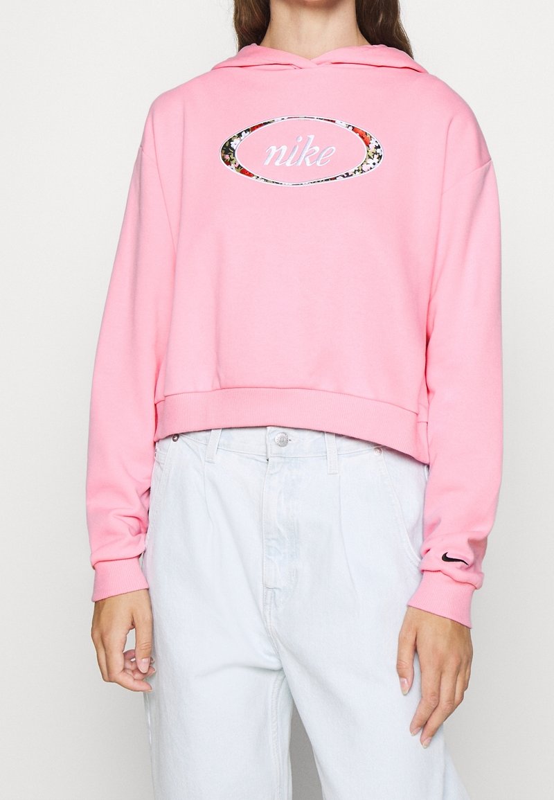 Rosa croppad hoodie i mjukt tyg med en blommig tryckt Nike-logotyp oval på bröstet och en liten svart swoosh på ärmen.
