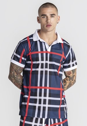 Młody mężczyzna o krótkich włosach i tatuażach, ubrany w białą koszulę w kratę w kolorach granatowym, czerwonym i białym z krótkim rękawem oraz pasujące krótkie spodenki.