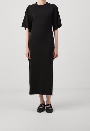 Persona que lleva un vestido negro largo con mangas cortas anchas y zapatos planos negros, de pie frente a un fondo claro y liso.