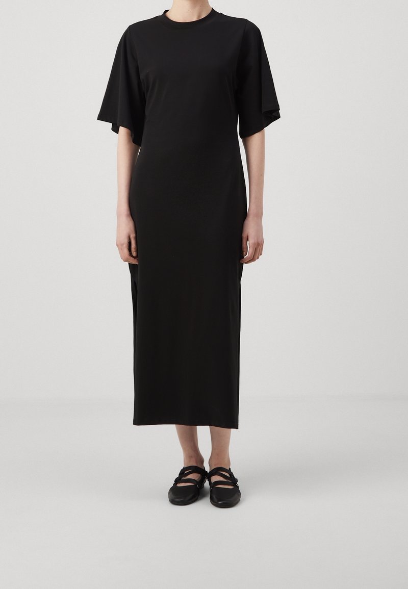 Personne portant une longue robe noire avec de larges manches courtes et des chaussures plates noires, debout devant un fond clair uni.