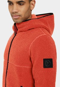 Fleece-ärmad jacka i klar orange med en texturerad yta. Har svarta dragkedjor och ett märke på vänster arm.