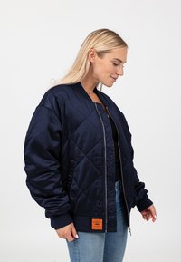 Navy gewatteerde bomberjack van gladde stof, met ribgebreide manchetten en kraag, voorin met rits, en een oranje label op de zoom.