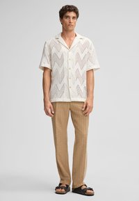 Korte mouwen, wit overhemd met knopen en een zigzagpatroon, gecombineerd met beige gestreepte broek en zwarte slidesandalen. Gestructureerde stof.