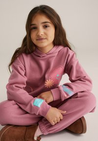 Lyserød hættetrøje med en smilende blomstergrafik, teksturerede detaljer på ærmerne, kombineret med matchende bukser og brune sko.