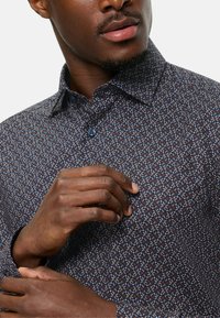 Camicia button-up di colore blu scuro con un motivo geometrico in blu chiaro, rosso e bianco. Presenta un colletto a punta e un dettaglio con un singolo bottone.