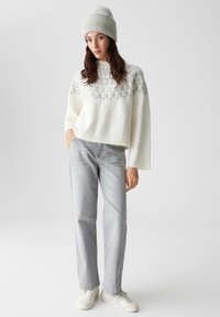 White knit sweater with gray pattern at the top, paired with light gray straight-leg jeans and white sneakers. Model wearing a matching gray beanie.