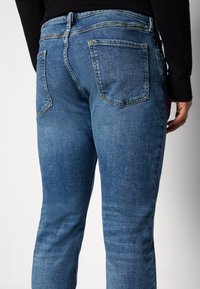 Jeans de mezclilla azul con un diseño ajustado, que cuentan con bolsillos traseros y detalles de costura visibles. La tela tiene una apariencia texturizada.