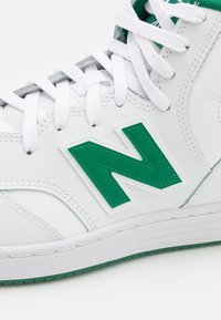 Vysoké tenisky s bílým koženým svrškem, zelenými akcenty a velkým zeleným logem "N". Mají texturovanou gumovou podrážku a bílé tkaničky.