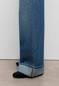 Donkerblauwe denim wijdvallende broek met rafelige zoom. De zoom is opgerold, waardoor een lichterblauwe binnenstof zichtbaar is. Zwart schoeisel is te zien aan de onderkant.