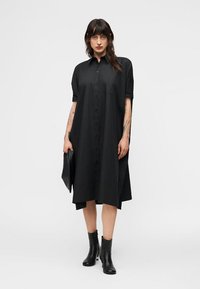 Γυναίκα που φοράει λασκώδη μαύρο φόρεμα μέσης μήκους με κουμπιά και κοντά μανίκια, κρατάει μαύρο τσαντάκι και φοράει μαύρα μποτάκια πάνω από τον αστράγαλο.